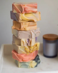 A stack of seven colorful Perennial Soaps Bar Soaps is arranged on a white surface, with a potted plant and a candle with a wooden lid in the background.