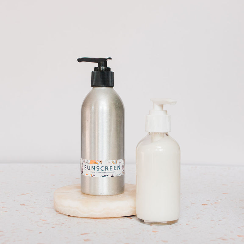 Two pump bottles of All Good Brand are side by side on a cream-colored circular surface: one labeled Sport Sunscreen Lotion SPF 30 and one clear with white lotion, set against a light background.