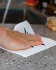 A hand wipes a gray speckled kitchen countertop with a Potluck Press Swedish Dishcloth. Sponges and a sink are visible in the background.